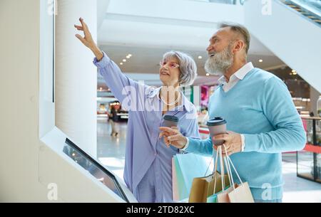 Aged smiley married couple standing at self-service kiosk in shopping mall Stock Photo
