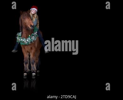 A young female equestrian on her festive decorated icelandic horse wearing a christmas wreath on black background, horse black shot Stock Photo