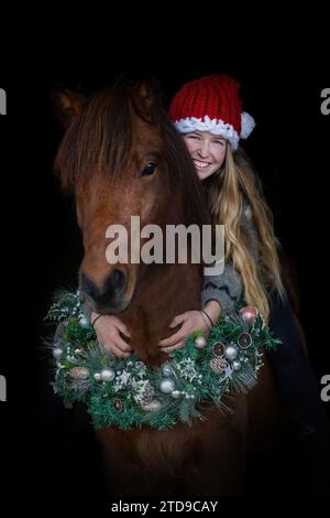 A young female equestrian on her festive decorated icelandic horse wearing a christmas wreath on black background, horse black shot Stock Photo