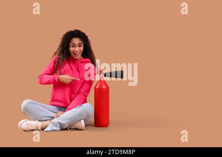 Young African-American woman pointing at fire extinguisher on brown background Stock Photo