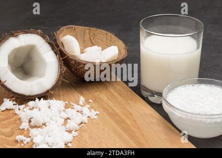 Coconut milk in glass and bowl. Coconut flakes, half fresh coconut, coconut shells on board. Black background. Close up Stock Photo