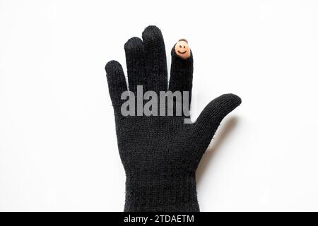 A black knitted glove with a cut finger and a cheerful smiley face is drawn on the finger, which is visible from the hole in the glove on a white back Stock Photo