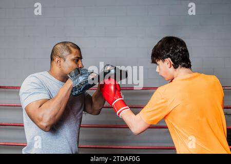 Horizontal photo caucasian teen boy wearing orange t-shirt and red boxing gloves, practicing boxing in the ring with his mid adult black trainer. Conc Stock Photo