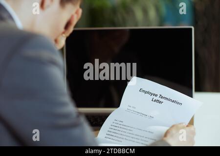 New job. Seen from behind modern middle aged woman employee in modern green office in grey business suit with laptop and employee termination letter. Stock Photo