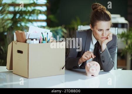 New job. pensive modern 40 years old woman employee in modern green office in grey business suit with personal belongings in cardboard box putting coi Stock Photo