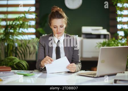 New job. pensive modern 40 years old woman employee in modern green office in grey business suit with laptop opening letter. Stock Photo