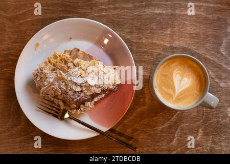 A latte in a coffee cup being drunk in the morning at a cafe next to an almond croissant. Heart symbol made in the cream on top of the coffee. UK Stock Photo