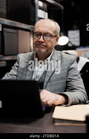 Elderly investigator checking confidential criminology evidence files on laptop. Private detective in agency file storage office surrounded by folders on cabinet shelves, portrait Stock Photo