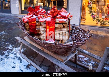 Snowy piles of firewood wrapped with red ribbons in a basket on a old wooden sledge in front oa shop as Christmas decoration in a mountain village Stock Photo