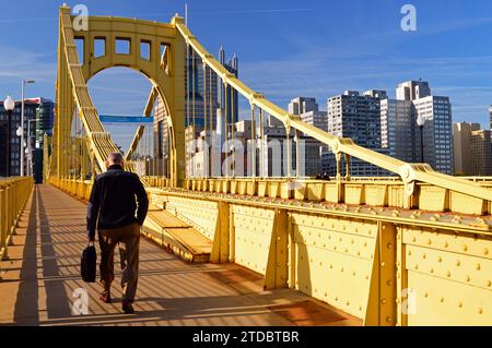 An adult man commutes to his job in downtown Pittsburgh Pennsylvania by walking across the yellow Roberto Clemente Bridge on a sunny day Stock Photo