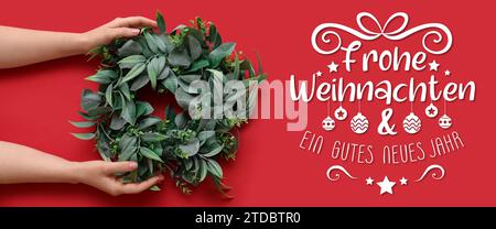 Text FROHE WEIHNACHTEN END GUTES NEUES JAHR (German for Merry Christmas and Happy New Year) and female hands with mistletoe wreath on red background Stock Photo