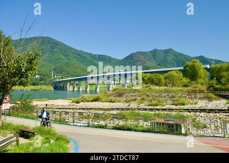 Hanam City, South Korea - October 1, 2023: A cyclist pedals along a bike path, with Deokpung Stream in the foreground and Paldang Bridge and Yebongsan Stock Photo