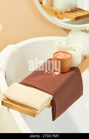 Close-up of a wooden tray placed across the bathtub with cloth towels, pink salt, scented candles and a bath sponge. Space with two main colors white Stock Photo