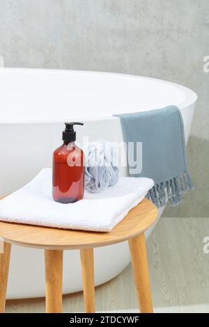 A bottle of brown shampoo and a bath sponge on a folded towel on a wooden table, next to a bathtub on a gray background. Space inside the bathroom for Stock Photo