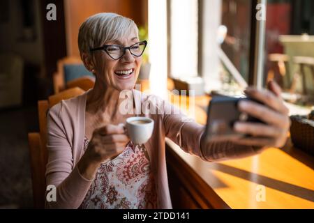 Mature woman is sitting in cafe and relaxing. She is using smartphone. Stock Photo