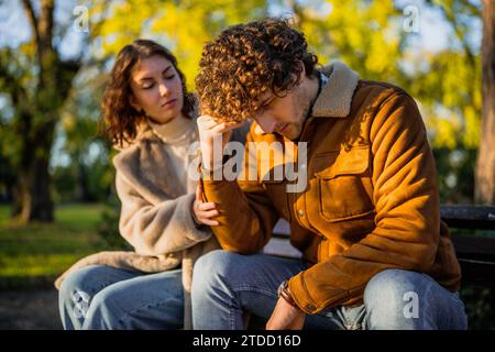 Young couple is sitting in park on sunny day. Man is sad and woman in consoling him. Stock Photo