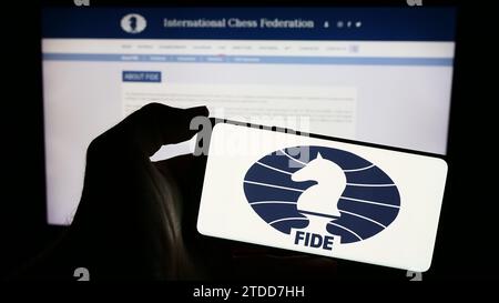 Person holding mobile phone with logo of organisation International Chess Federation (FIDE) in front of web page. Focus on phone display. Stock Photo