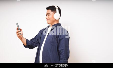 Happy young Asian man wearing wireless headphones listening to music from smartphone studio shot isolated on white background Stock Photo