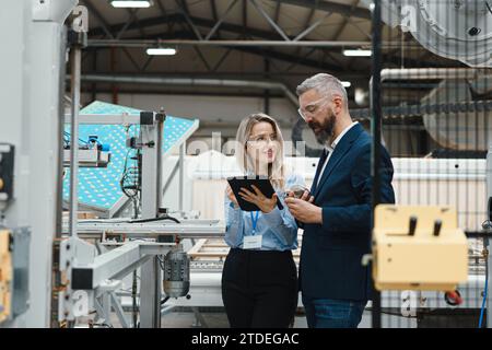 Female engineer and male project manager standing in modern industrial factory by precision robotic arm. Robot assembling wooden furniture in big Stock Photo