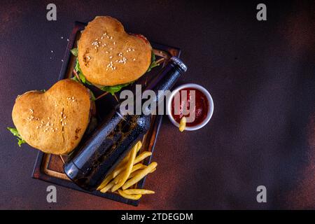 Valentine day Heart-shaped burgers. Two tasty cheeseburgers with french fries and beer bottles on dark table background. Idea for Valentine day dinner Stock Photo