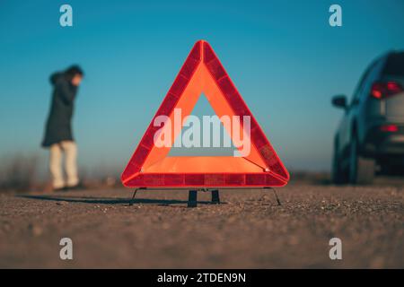 Vehicle breakdown, red warning triangle on the road with a broken down car and woman making a phone call to road service, selective focus Stock Photo