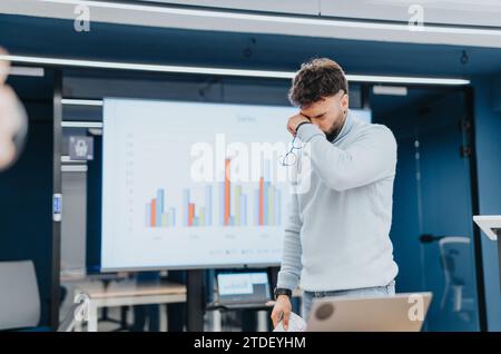 A young, tired businessperson rubs his eyes after working the entire day on the project in the office. Stock Photo