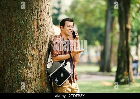 Smiling young man leaning on tree when talking on phone with friend Stock Photo