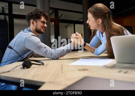 Side view of joking male and female colleagues doing arm wrestling at desk Stock Photo
