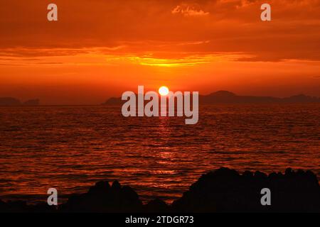 Vibrant red sunset over the ocean with sun setting behind island silhouettes and rocky shore in Thailand Stock Photo
