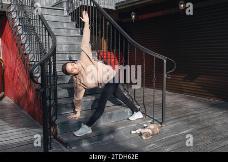 A young woman plays sports on the street near a street cat, performs Trikonasana exercise, triangle pose, practices yoga in sportswear standing on the Stock Photo