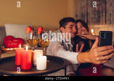 Valentines Day loving couple taking selfie on phone by wine glasses with candles having pizza romantic dinner at home. Happy man and woman celebrating Stock Photo