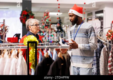 Worker helping elderly woman to choose right fit of formal attire, preparing to attend festive dinner on christmas holiday. African american store employee recommending clothes for client. Stock Photo