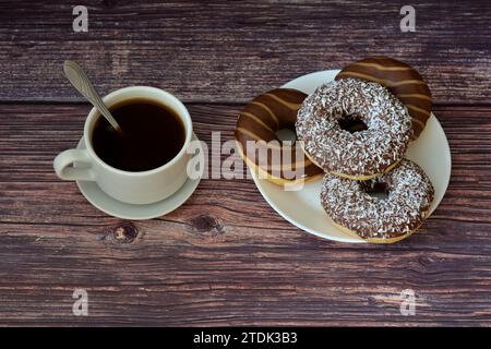 A cup of hot black coffee on a saucer with a spoon and a plate with several chocolate-coated donuts on a wooden table. Top view, flat lay. Stock Photo