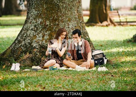 Positive young couple resting under big tree in park, having snacks and discussing posts on social media Stock Photo