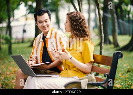 University students sitting on bench in park and discussing project they are working on together Stock Photo