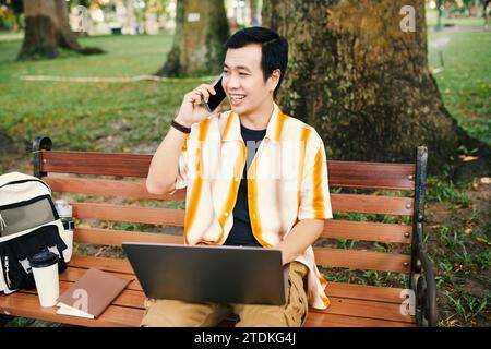 Portrait of smiling Vietnamese man sitting on bench in park, working on laptop and talking on phone Stock Photo