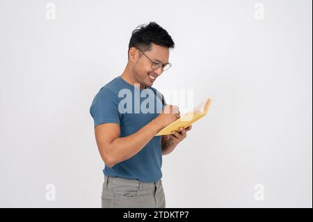 A positive Asian man in casual clothes is keeping his diary, writing something in a book while standing on an isolated white studio background. Stock Photo