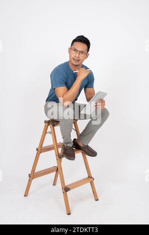 A stressed, serious Asian man in casual clothes is sitting on a wooden ladder with a digital tablet in his hand, isolated white background. People and Stock Photo