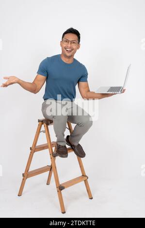 A cheerful and excited Asian man in casual clothes is sitting on a wooden ladder with a laptop computer in his hand, showing extremely happy expressio Stock Photo