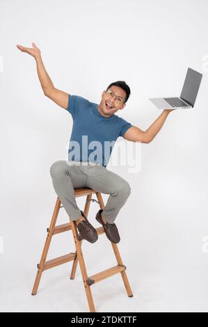 A cheerful and excited Asian man in casual clothes is sitting on a wooden ladder with a laptop computer in his hand, showing extremely happy expressio Stock Photo