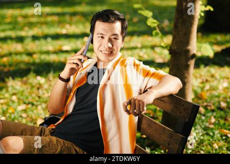 Cheerful young Asian man sitting on bench in park and calling on phone Stock Photo