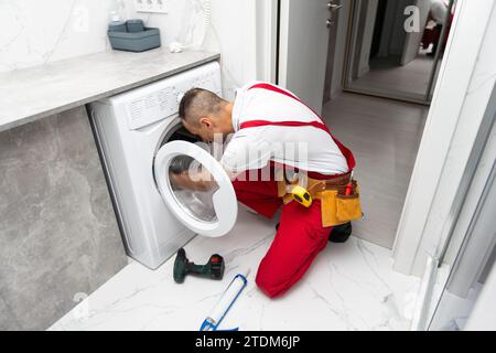 Young smiling happy man plumber handyman in overalls repair fix the washing machine in the kitchen and look to the camera Stock Photo