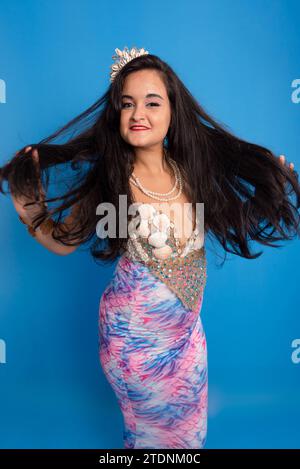 Beautiful woman, wearing a tiara and seashell bracelets, against a blue background, touching her black hair. Yemanja worshiper. Stock Photo