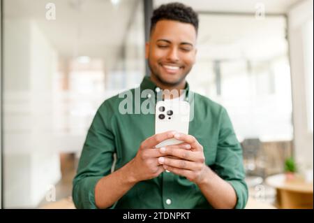 Selective focus on the smartphone in hands of happy and cheerful indian man, male office employee using phone, scrolling news feed in social medias, chatting online, using mobile app Stock Photo