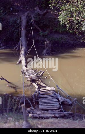 A homemade wooden bridge built over a river with railings made of ropes. Boards nailed to base of fallen tree trunk in water Stock Photo