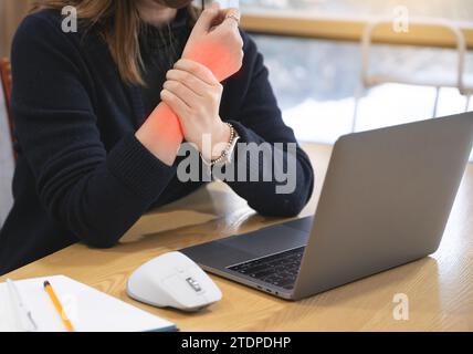A woman working at a desk using a laptop and a mouse is suffering from wrist pain, and the painful part is marked in red Stock Photo
