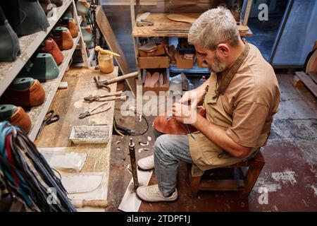 High angle of mature experienced shoemaking master in casualwear sitting in front of table and shelves with footwear workpieces Stock Photo
