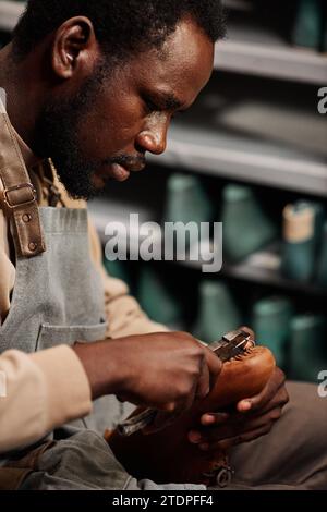 Side view of young African American man in apron creating new boot or shoe in workshop and fixing together upper part and sole Stock Photo