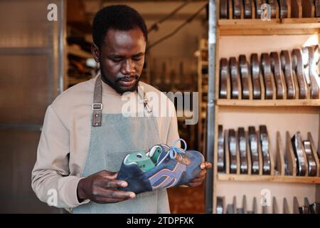 Young black man in workwear looking at workpiece of grey suede sports shoe in his hands while standing against shelves in workshop Stock Photo