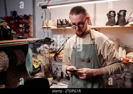 Young serious man in grey apron texting in mobile phone while standing in front of camera in workshop and communicating with online clients Stock Photo